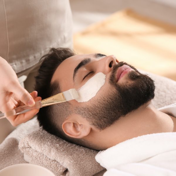 Cosmetologist applying mask on man's face in spa salon